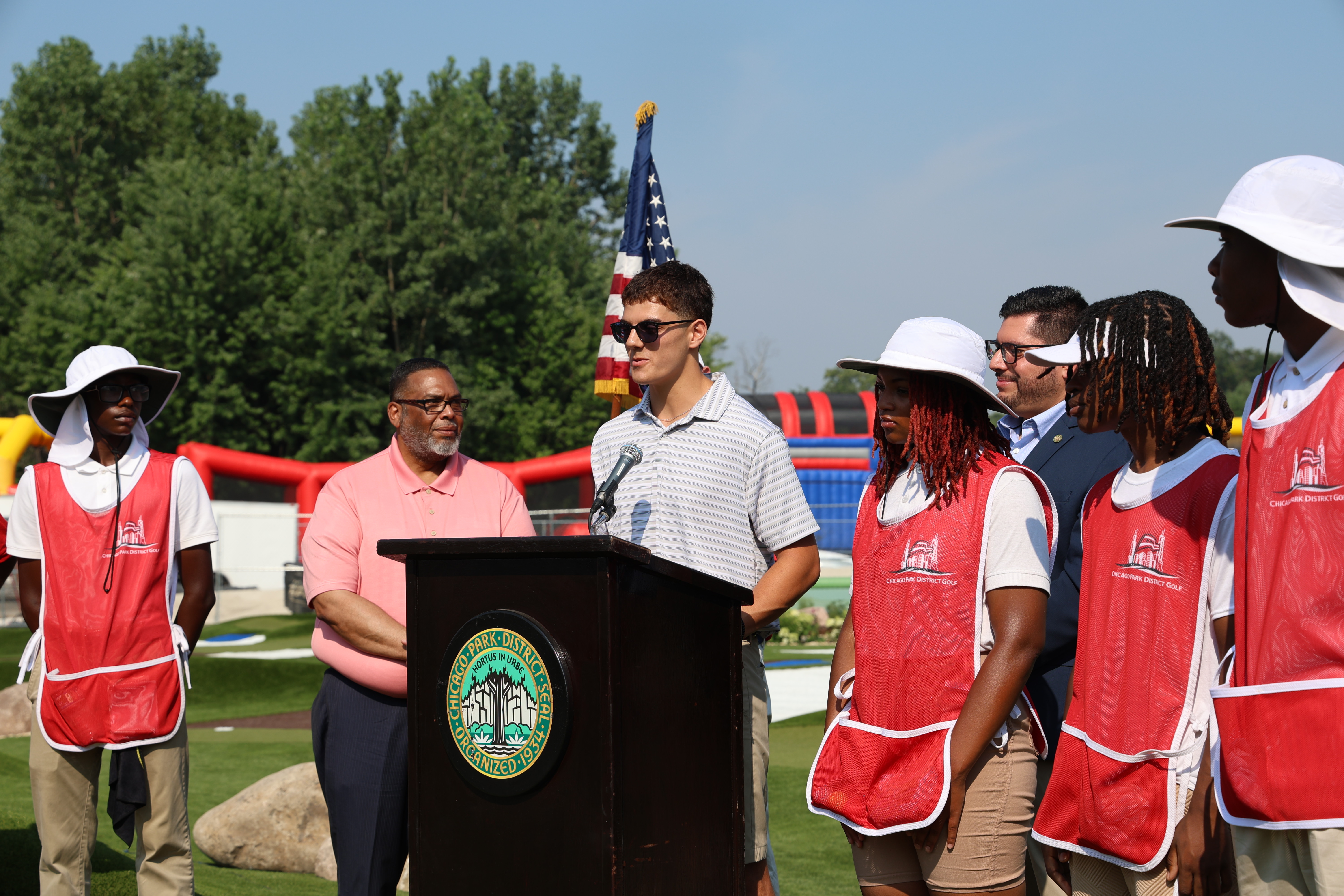 A man speaks at a podium with a Chicago Park District logo, flanked by staff in red vests. An American flag stands nearby.